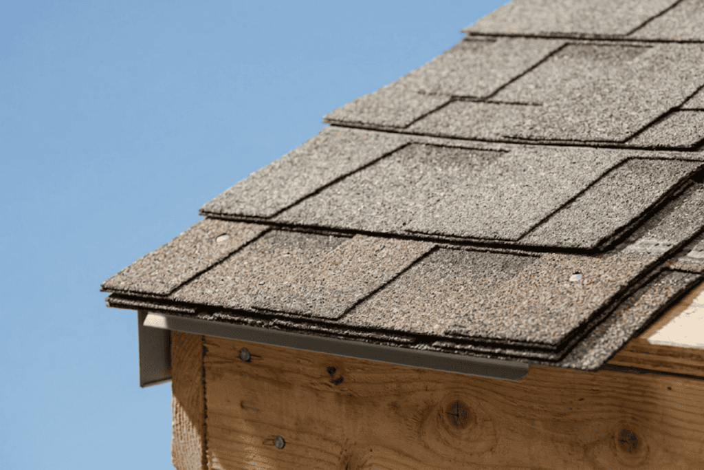 Close-up of gray asphalt shingles stacked on a wooden roof overhang against a blue sky.