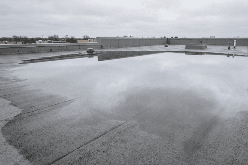 Flat commercial roof with a large puddle, reflecting the cloudy sky and a low edge wall with vents along the perimeter.